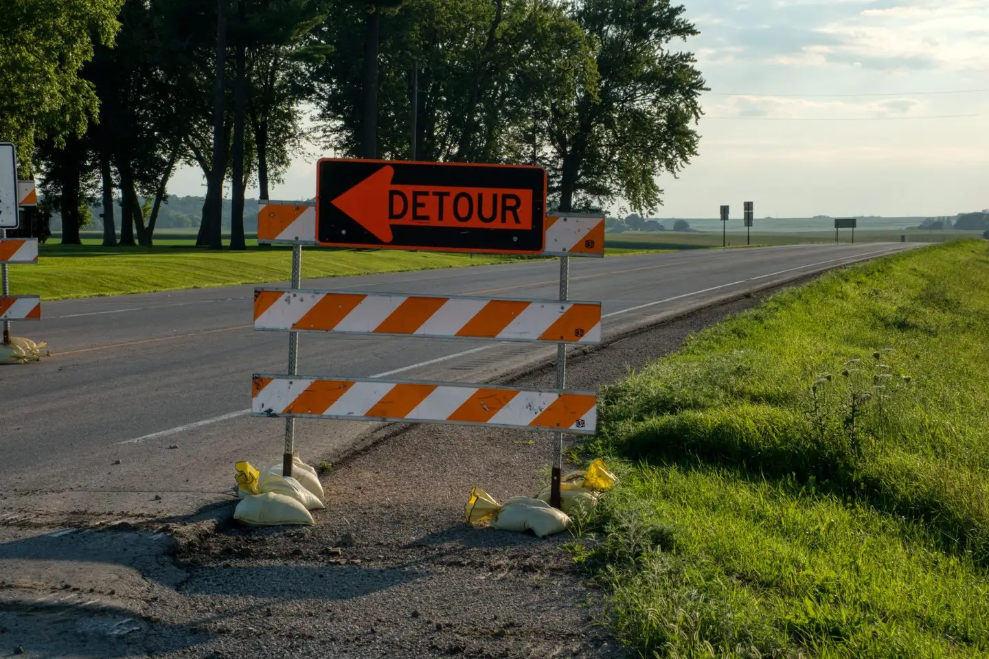 An orange detour sign with an arrow on a striped road barrier beside a rural road.